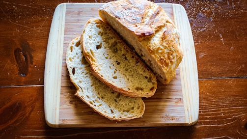 A loaf of rustic bread with a golden crust sits on a rectangular wooden cutting board. Two slices are cut from the loaf, displaying a soft and airy interior. The cutting board is placed on a rich brown wooden table, providing a warm and natural background.