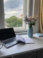 A serene workspace with a laptop, notebook, and a cup of tea beside a window with soft natural light.