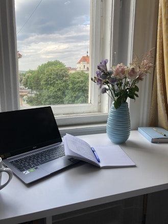 A calm and serene workspace featuring a white desk near a window with a view of lush trees and a building with a red roof. On the desk, there is an open laptop, a notebook with a pen resting on it, a vase with a bouquet of pastel-colored flowers, and a cup. The window allows natural light to brighten the space.