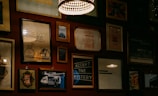 A wall adorned with various framed artworks and photographs, including images of a bicycle, inspirational quotes, and abstract designs. The lighting is warm and subtle, with a prominent hanging lamp creating a cozy atmosphere.