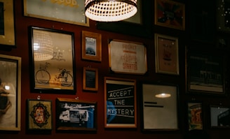A wall adorned with various framed artworks and photographs, including images of a bicycle, inspirational quotes, and abstract designs. The lighting is warm and subtle, with a prominent hanging lamp creating a cozy atmosphere.