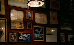 A wall adorned with various framed artworks and photographs, including images of a bicycle, inspirational quotes, and abstract designs. The lighting is warm and subtle, with a prominent hanging lamp creating a cozy atmosphere.