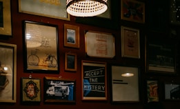 A wall adorned with various framed artworks and photographs, including images of a bicycle, inspirational quotes, and abstract designs. The lighting is warm and subtle, with a prominent hanging lamp creating a cozy atmosphere.