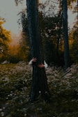 In a dense forest with abundant greenery, a pair of hands, wearing a white shirt, is seen hugging a tree trunk. The background features dappled sunlight filtering through the tall trees and a ground covered in wild plants and dandelions.