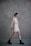 A person in a light-colored outfit walks against a textured gray wall, showcasing a relaxed and stylish fashion choice with a combination of a bralette, shorts, and an open shirt. Black combat boots complement the casual look. The hair is styled in a bun, adding to the polished appearance.