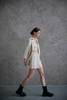 A person in a light-colored outfit walks against a textured gray wall, showcasing a relaxed and stylish fashion choice with a combination of a bralette, shorts, and an open shirt. Black combat boots complement the casual look. The hair is styled in a bun, adding to the polished appearance.