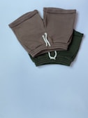 Two pairs of shorts, one in a brownish-grey color and the other in dark green, are placed overlapping on a white surface. Both have white drawstrings and elastic waistbands.