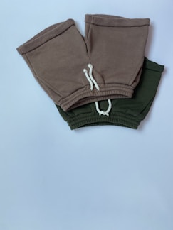 Two pairs of shorts, one in a brownish-grey color and the other in dark green, are placed overlapping on a white surface. Both have white drawstrings and elastic waistbands.