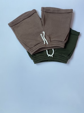 Two pairs of shorts, one in a brownish-grey color and the other in dark green, are placed overlapping on a white surface. Both have white drawstrings and elastic waistbands.