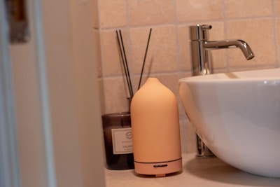 A beige ceramic aroma diffuser sits on a bathroom counter next to a dark brown candle jar labeled with text and a white ceramic vessel sink. The background features beige tiled walls.