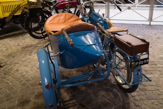A vintage motorcycle with a sidecar painted in blue is parked on a stone-paved surface. The sidecar has a tan leather seat, and there is a brown wooden box mounted on the rear of the motorcycle. The license plate reads 'Y-111-YP'. Several other vintage vehicles are visible in the background.
