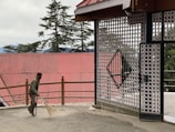 A person is sweeping an outdoor area near a decorative metal gate with circular designs. The background features tall evergreen trees and a building with a red corrugated roof. The place appears to be peaceful and somewhat rustic.