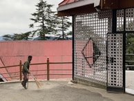 A person is sweeping an outdoor area near a decorative metal gate with circular designs. The background features tall evergreen trees and a building with a red corrugated roof. The place appears to be peaceful and somewhat rustic.
