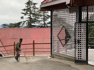 A person is sweeping an outdoor area near a decorative metal gate with circular designs. The background features tall evergreen trees and a building with a red corrugated roof. The place appears to be peaceful and somewhat rustic.