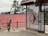 A person is sweeping an outdoor area near a decorative metal gate with circular designs. The background features tall evergreen trees and a building with a red corrugated roof. The place appears to be peaceful and somewhat rustic.