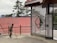 A person is sweeping an outdoor area near a decorative metal gate with circular designs. The background features tall evergreen trees and a building with a red corrugated roof. The place appears to be peaceful and somewhat rustic.