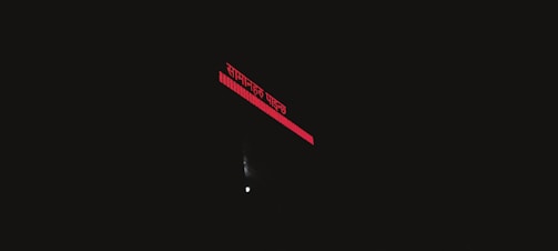 A dark image with a bright red illuminated sign showing text in a script. Below the sign, a faint light source is visible, possibly from a small window or light bulb.