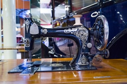 A vintage sewing machine with intricate details and designs on its body is situated on a wooden table. Its metallic surface reflects light, showcasing a combination of dark and shiny textures. A wheel and a spool holder are part of its robust and classic design. The background includes blurred elements that suggest an indoor setting with various colors.