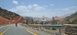 A scenic road runs through a mountainous landscape with a mixture of red earth and green vegetation. A motorcycle is traveling on the paved road, which is marked by yellow lines. A triangular road sign warns of a winding road ahead. The sky is partly cloudy, and the distant mountains provide a picturesque backdrop.