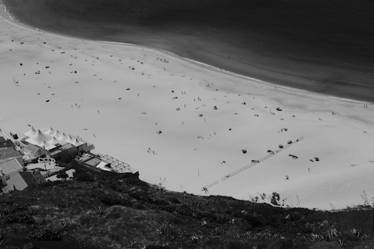 An aerial view of a beach with scattered people and umbrellas. The sand appears light and the sea is a dark contrast. A structure with white tents is visible near the bottom left corner, surrounded by buildings and vegetation.