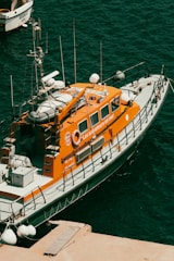 A bright orange rescue boat is moored at a dock, featuring several antennas and life-saving equipment. The boat's side is marked with the phrase 'Les sauveteurs en mer'. The surrounding water is a deep blue-green color, with another small boat visible partially in the background.