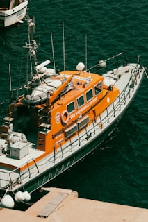 A bright orange rescue boat is moored at a dock, featuring several antennas and life-saving equipment. The boat's side is marked with the phrase 'Les sauveteurs en mer'. The surrounding water is a deep blue-green color, with another small boat visible partially in the background.