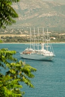 A large white cruise ship with several masts is sailing on a calm blue sea, partially framed by green foliage. In the background, a rocky landscape and a shoreline with scattered houses are visible, under a partially cloudy sky.