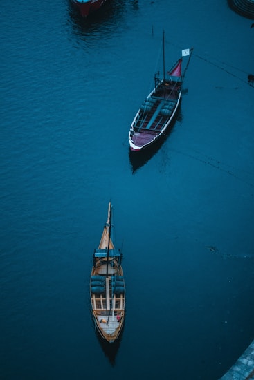 Two wooden boats float on a deep blue expanse of water, with one boat positioned closer to the viewer than the other. The boats feature traditional designs, with the larger one displaying a flag and some barrels on deck, while the smaller one appears empty.