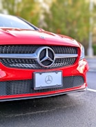 A close-up of the front section of a red Mercedes-Benz car, focusing on the grille and emblem. The Mercedes-Benz logo is prominently displayed in the center. The car is parked on a roadway with a blurred background of greenery.