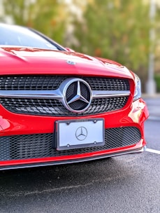 A close-up of the front section of a red Mercedes-Benz car, focusing on the grille and emblem. The Mercedes-Benz logo is prominently displayed in the center. The car is parked on a roadway with a blurred background of greenery.