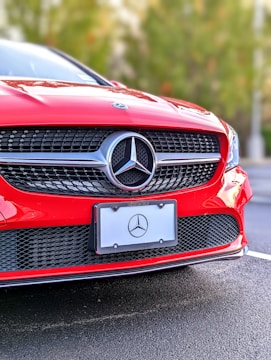 A close-up of the front section of a red Mercedes-Benz car, focusing on the grille and emblem. The Mercedes-Benz logo is prominently displayed in the center. The car is parked on a roadway with a blurred background of greenery.