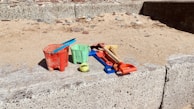 Colorful plastic beach toys, including red and green buckets, a blue spade, red and yellow rakes, and a tennis ball, are arranged on concrete beside a sandy area with scattered pebbles.