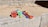 Colorful plastic beach toys, including red and green buckets, a blue spade, red and yellow rakes, and a tennis ball, are arranged on concrete beside a sandy area with scattered pebbles.
