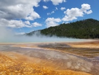 Geothermal hot spring with vibrant, mineral-rich terraces. Steam rises above the spring, contrasting with a backdrop of lush, forested hills under a partly cloudy blue sky.