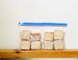 Slices of bread wrapped in plastic are neatly placed inside two resealable bags, resting on a wooden surface against a plain white background.