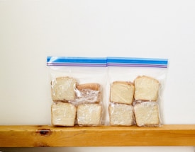 Slices of bread wrapped in plastic are neatly placed inside two resealable bags, resting on a wooden surface against a plain white background.