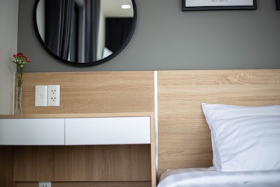 A modern bedroom scene features a wooden headboard and a sleek nightstand with a single drawer. A round black-framed mirror is mounted on the wall above. A white pillow with subtle striped patterns rests on the bed. A vase with red flowers is placed on the nightstand next to a power outlet.
