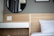 A modern bedroom scene features a wooden headboard and a sleek nightstand with a single drawer. A round black-framed mirror is mounted on the wall above. A white pillow with subtle striped patterns rests on the bed. A vase with red flowers is placed on the nightstand next to a power outlet.