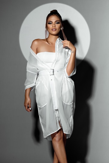 A confident young woman wearing a chic beige dress standing against a soft cream background.