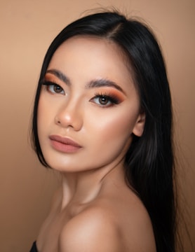 A portrait of a person with long black hair and brown eyes. The person has smooth skin, orange and brown eye makeup, and is wearing a neutral lip color. The background is a solid beige color.