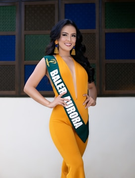 A woman is posing confidently, wearing a vibrant yellow jumpsuit with a deep V-neckline and a sash that reads 'BALER AURORA.' She has long, styled black hair and is accessorized with large tassel earrings. The background consists of a geometric pattern of colored glass panes in shades of green, blue, and brown.
