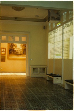 A room with a patterned tiled floor and beige walls. On one wall, there's a projection of French text from an old newspaper, while another wall displays framed artwork, possibly a historic photograph or painting. The ceiling has a light fixture, and benches are placed along the walls.
