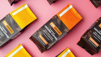 Multiple packages of mushroom-based ground coffee are displayed on a pink surface. The packaging is primarily brown with yellow-orange accents, featuring the brand name PureShrooms prominently. Text on the packages indicates that the coffee includes mushrooms like chaga and cordyceps and is labeled as a medium roast.