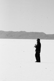 A solitary figure dressed in dark clothing and a hat stands in the vast expanse of a bright, flat landscape, holding and playing a flute. The background is a range of distant mountains under an overcast sky.