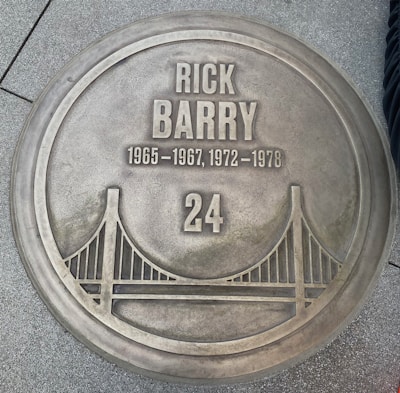 A metallic circular plaque on the ground featuring the name Rick Barry, with years 1965-1967 and 1972-1978 inscribed below. The number 24 is prominently displayed at the bottom, accompanied by an image of a bridge.