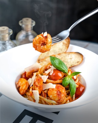 A plate of pasta topped with shrimp, cherry tomatoes, and grated cheese, garnished with fresh basil leaves. Slices of toasted bread are placed behind the pasta. A fork holds a shrimp above the plate, with steam rising from it, suggesting that the dish is hot.