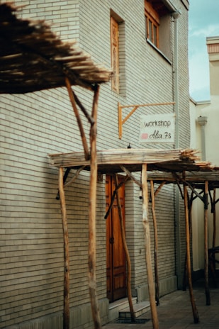 A narrow alley features a beige brick building with a wooden door. Above the door, a small sign reads 'workshop Alia 7s' indicating a creative or artisanal space. The structure is supported by wooden beams that form a rustic pergola over part of the pathway. The scene feels quiet and secluded.