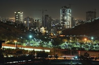 A nighttime urban landscape featuring tall buildings illuminated by lights, with cranes visible on the rooftops indicating ongoing construction. There is a well-lit highway in the foreground with light trails of moving vehicles, and green spaces with street lights scattered among trees.