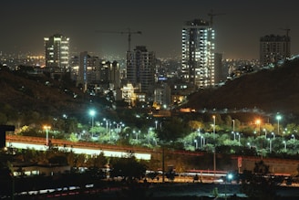 A nighttime urban landscape featuring tall buildings illuminated by lights, with cranes visible on the rooftops indicating ongoing construction. There is a well-lit highway in the foreground with light trails of moving vehicles, and green spaces with street lights scattered among trees.