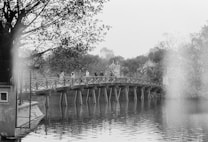 A black and white image depicting a wooden bridge spanning over a calm body of water. Trees with lush foliage frame the scene on both sides. Several people are walking across the bridge, and in the background, buildings are partially visible through a slight haze.